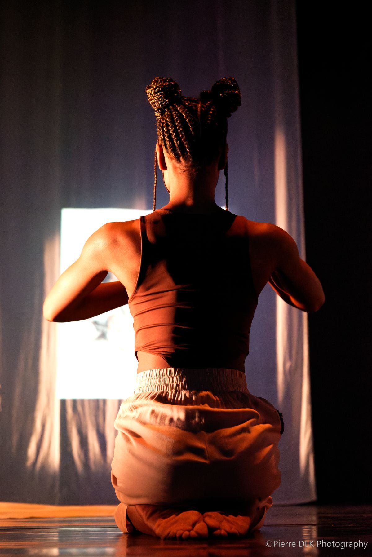 Massinda Zinga kneeling on stage, her back to the audience, in warm stage light