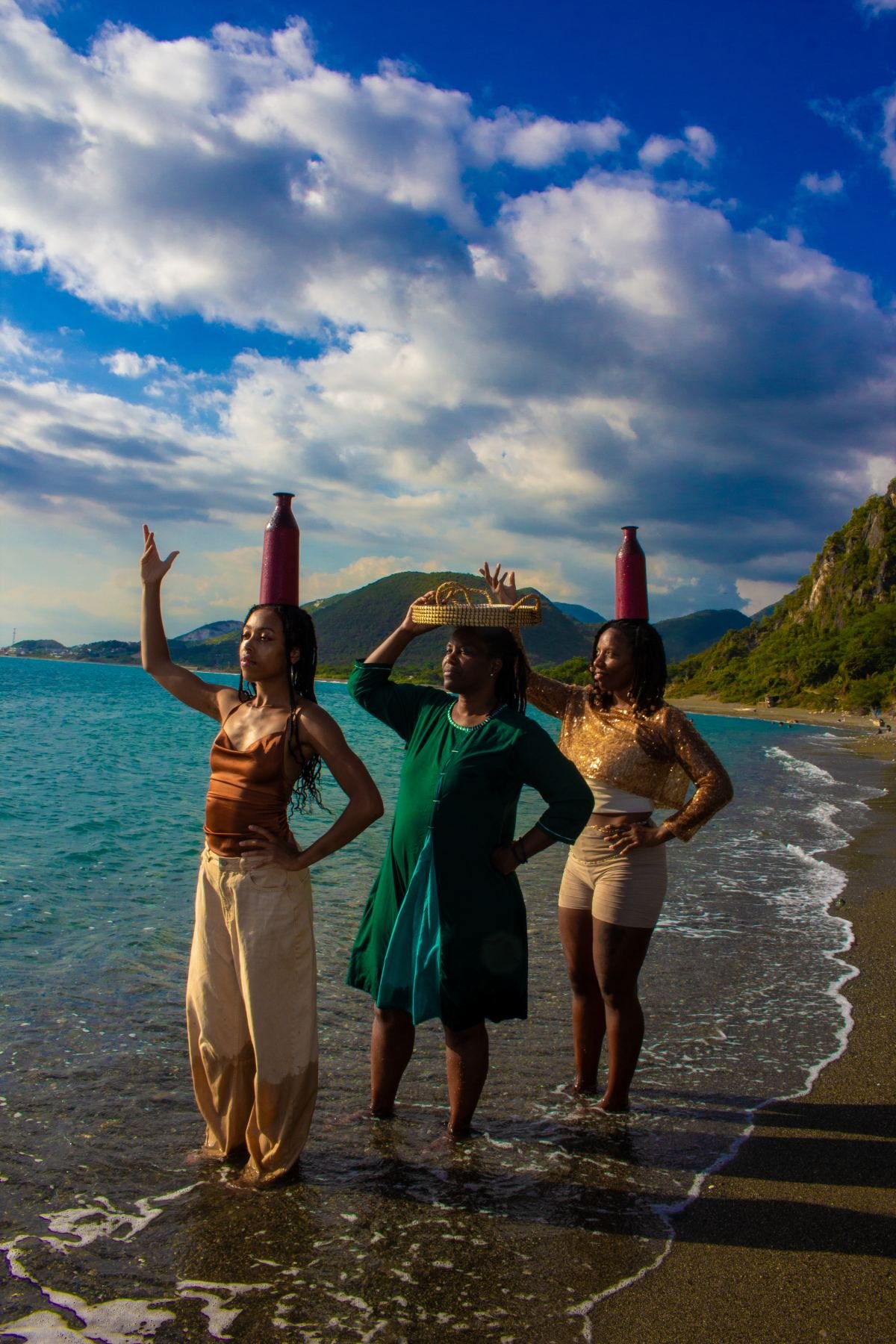 Three women standing in the sea on a Jamaican shore, two balancing tall red vessels on their heads