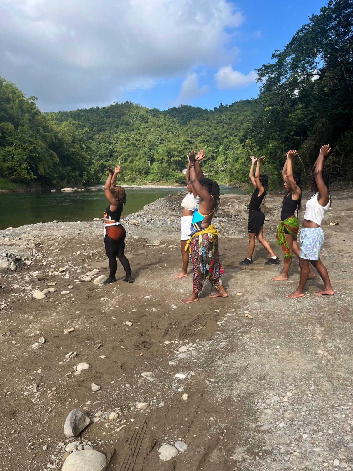 Women dancing on a riverbank surrounded by green hills