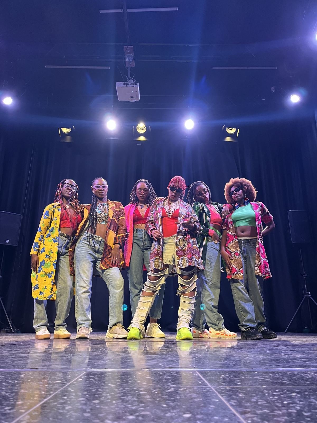 Six performers in colourful coordinated outfits on a stage under spotlights