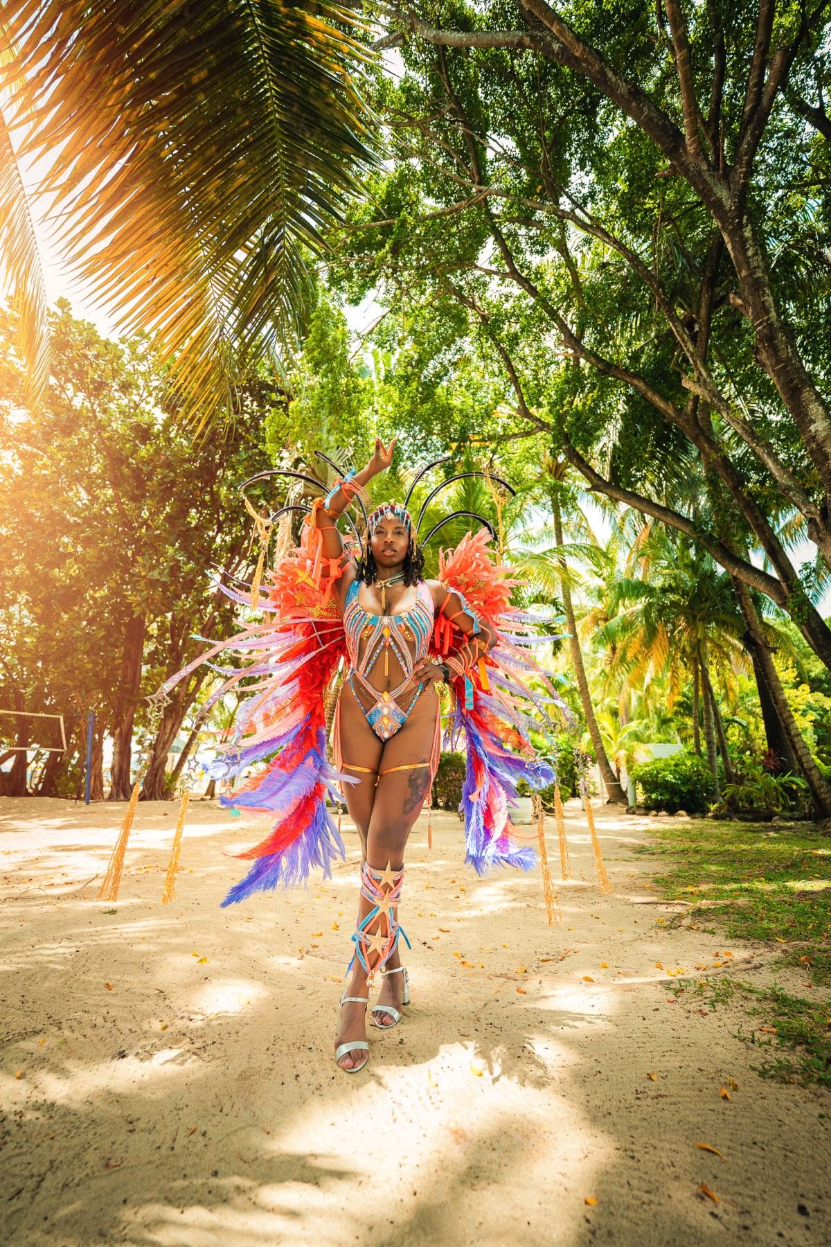 Massinda Zinga in a feathered carnival costume on a sandy path under palm trees, one arm raised