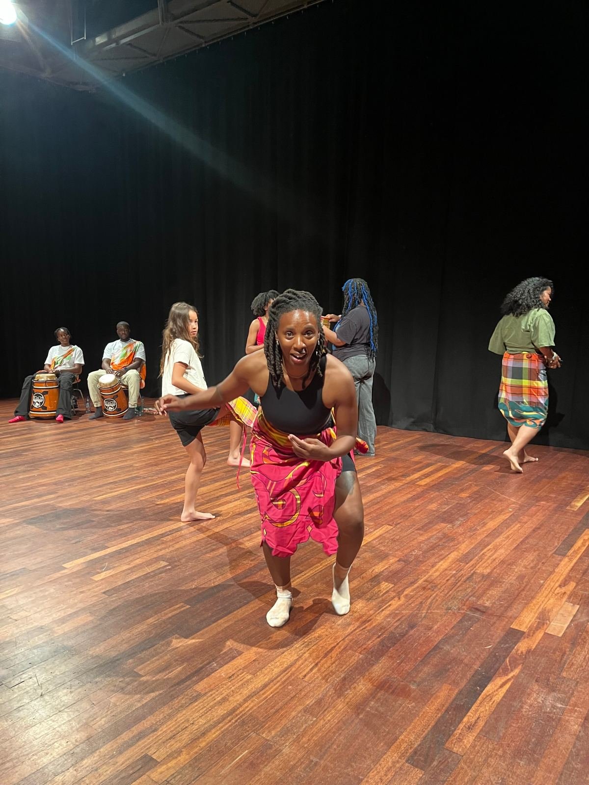 Massinda Zinga dancing in a studio in a pink wrap skirt, knees bent, with workshop participants and two drummers behind her