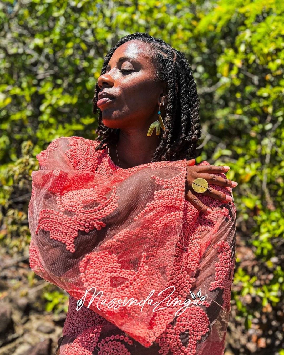 Massinda Zinga in a coral sequined lace top, eyes closed, one hand on her shoulder, against green foliage