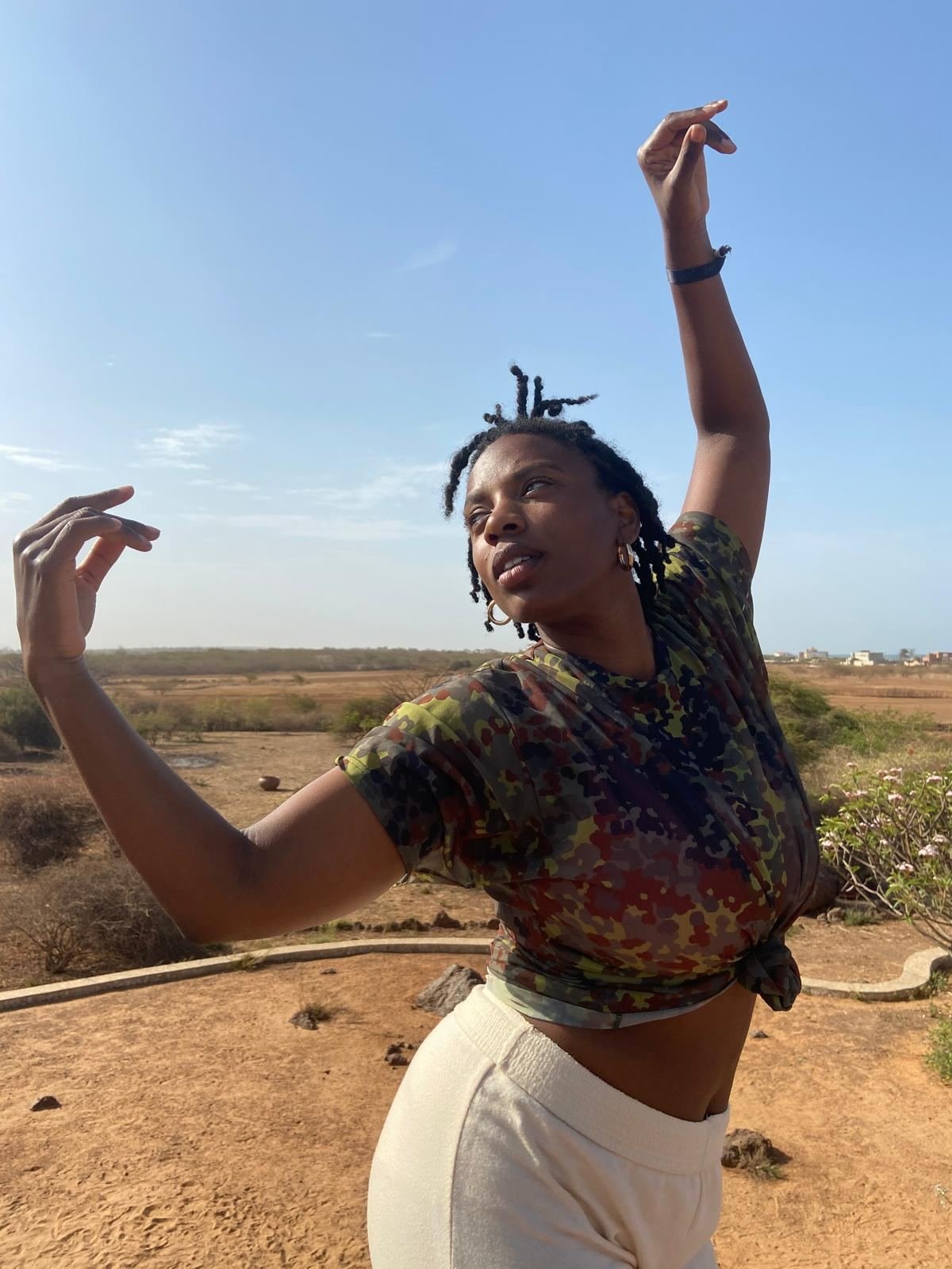 A woman dancing outdoors under a blue sky, both arms raised, in a camouflage-print T-shirt