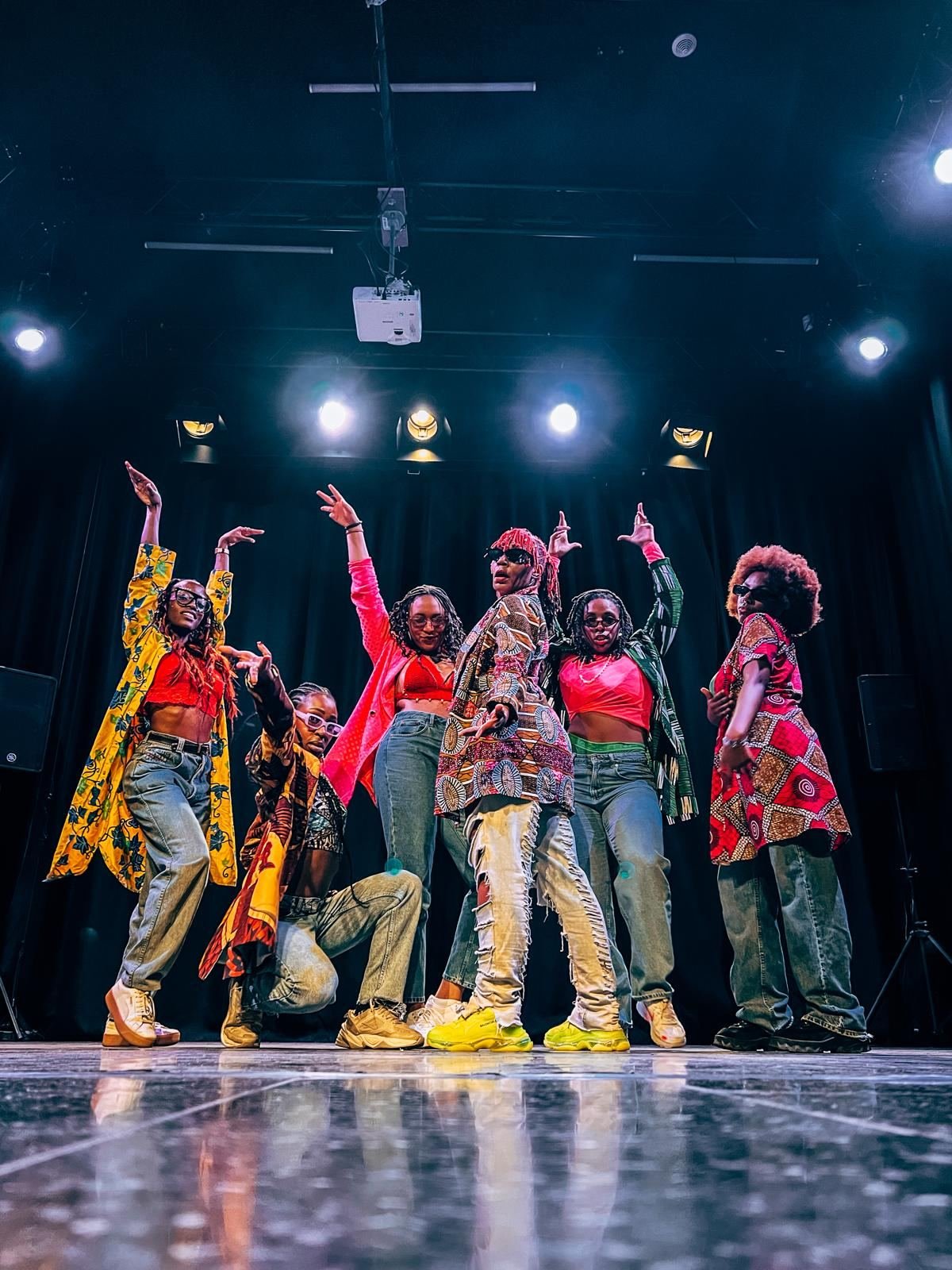 Six dancers in African-print outfits and jeans striking a pose on stage under the lights, arms raised