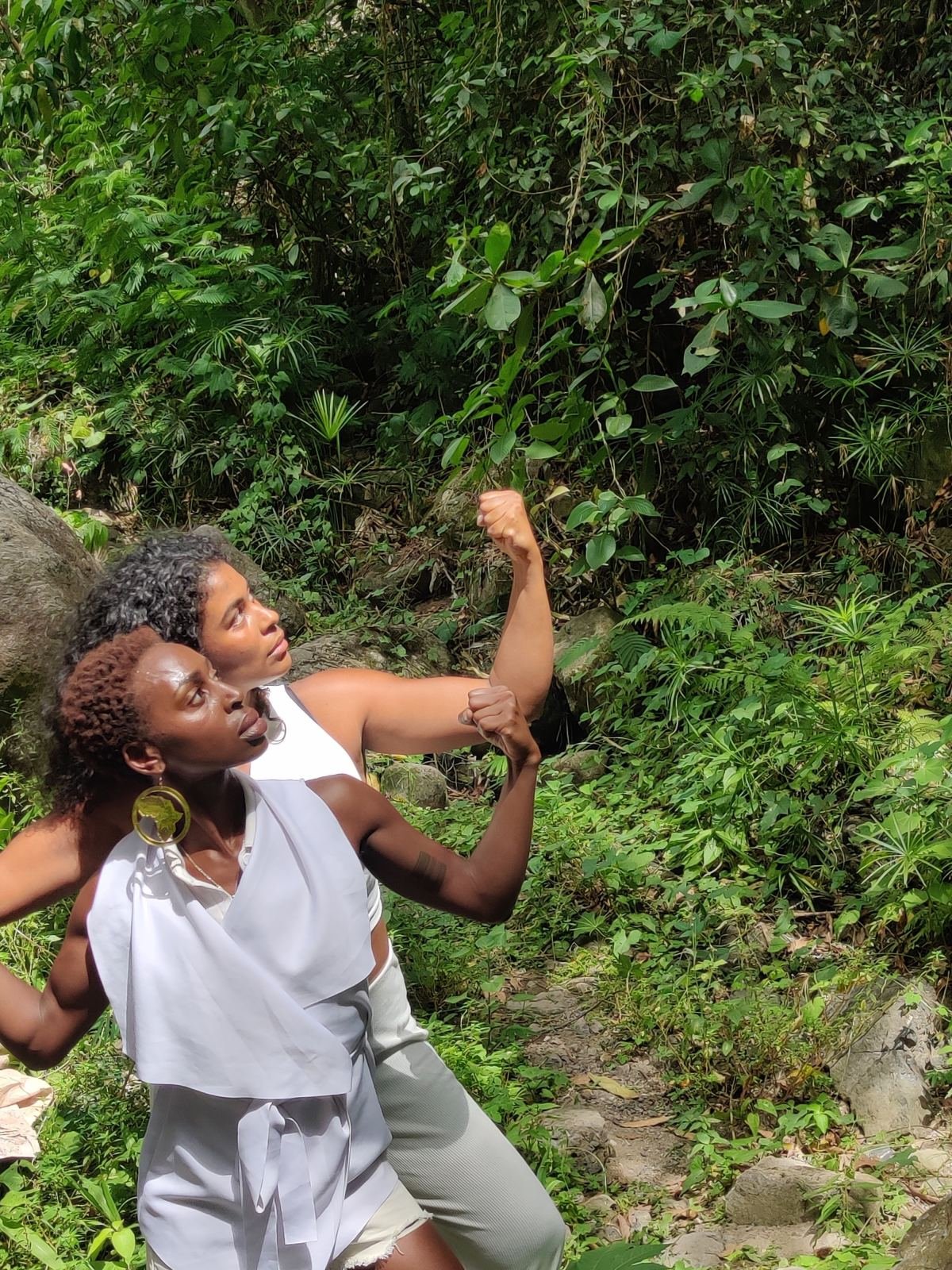 Two dancers dressed in white in a tropical forest, each flexing an arm and looking up