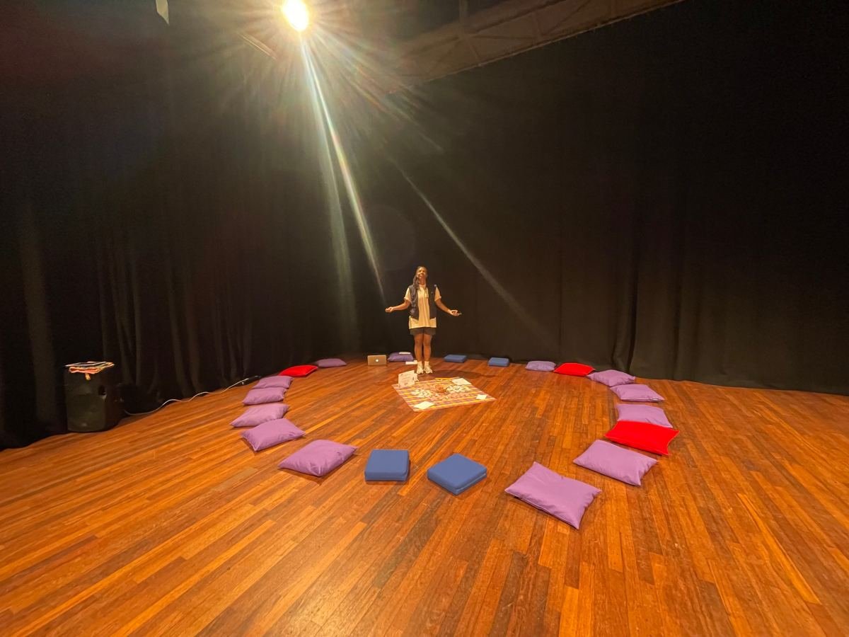 Massinda Zinga standing in a beam of light in a dark studio, at the head of a circle of cushions laid around a madras cloth