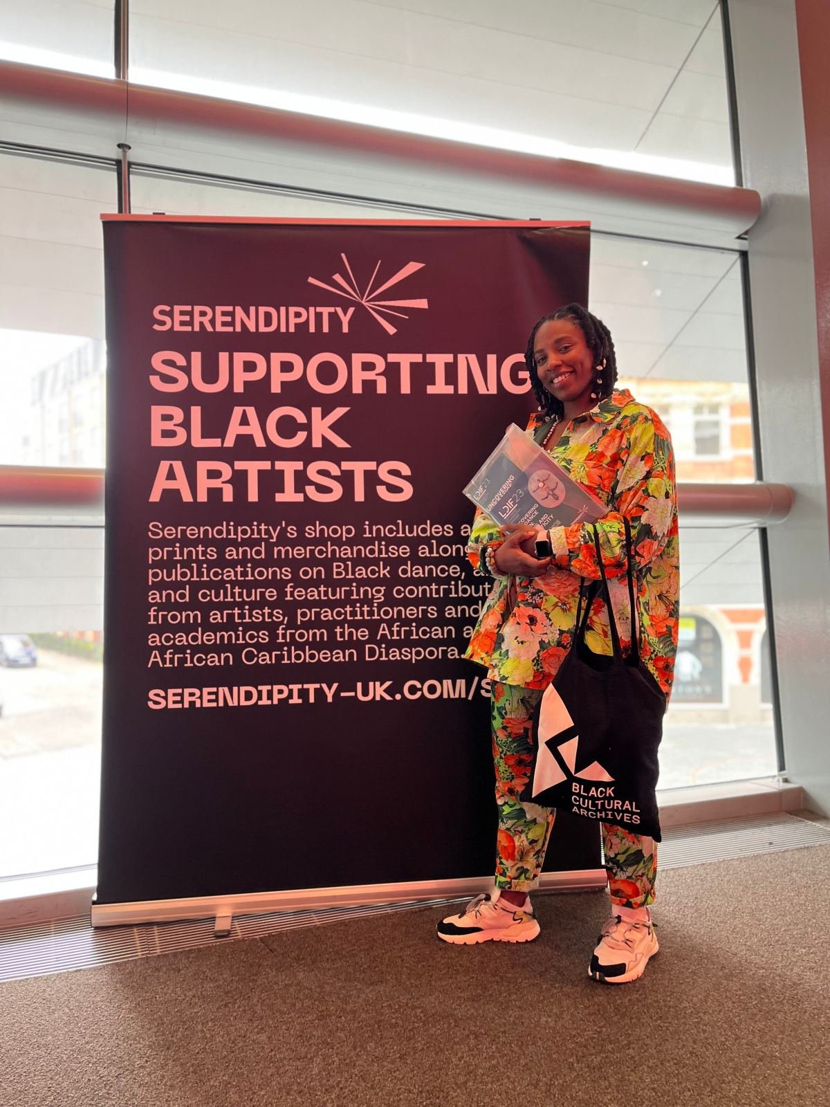 Massinda Zinga in a floral suit beside a Serendipity banner supporting Black artists, holding a folder and a Black Cultural Archives tote bag