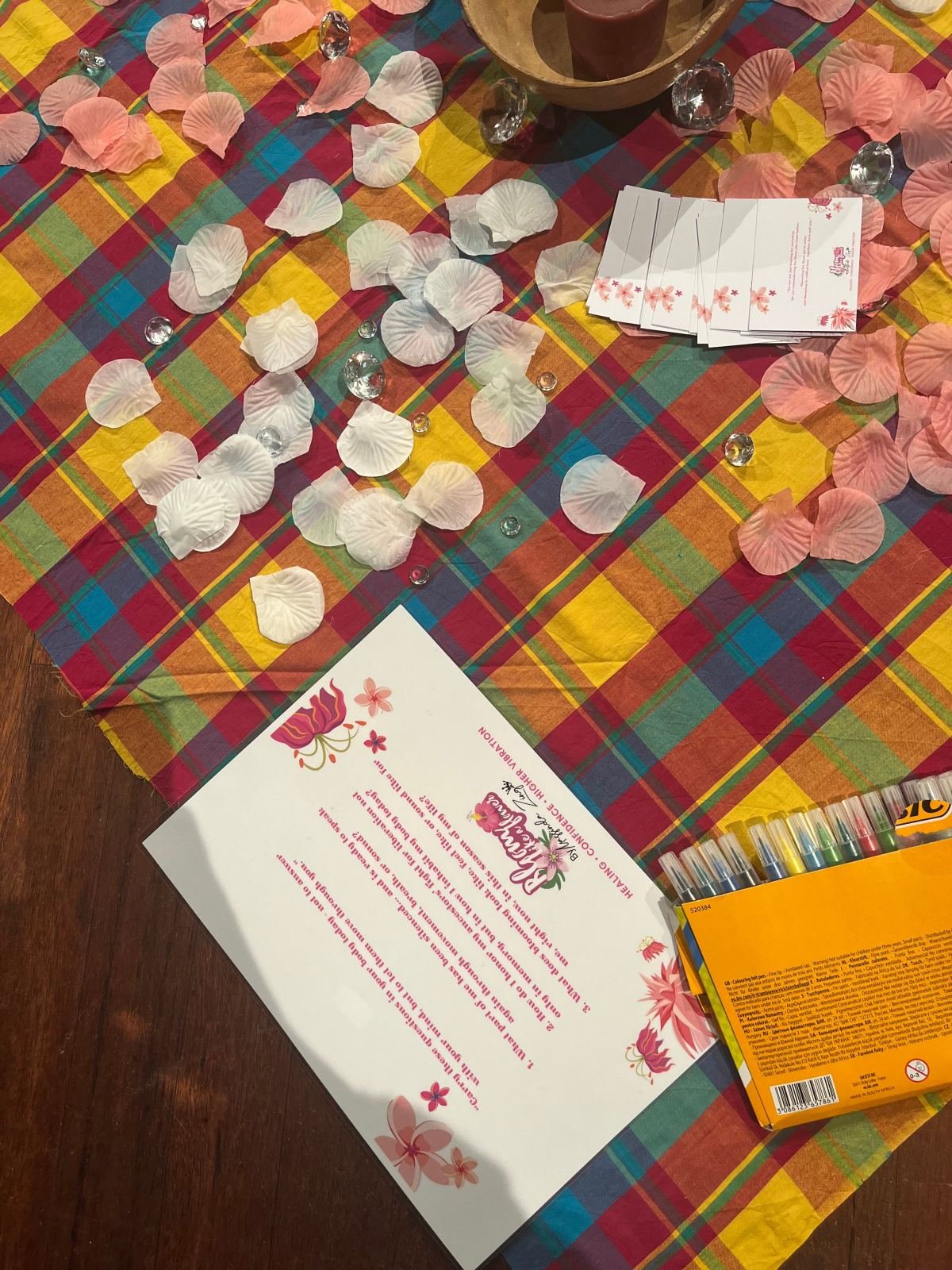Des pétales, des cristaux et des cartes sur un tissu madras, avec une fiche Bloom Like a Flower et une boîte de feutres