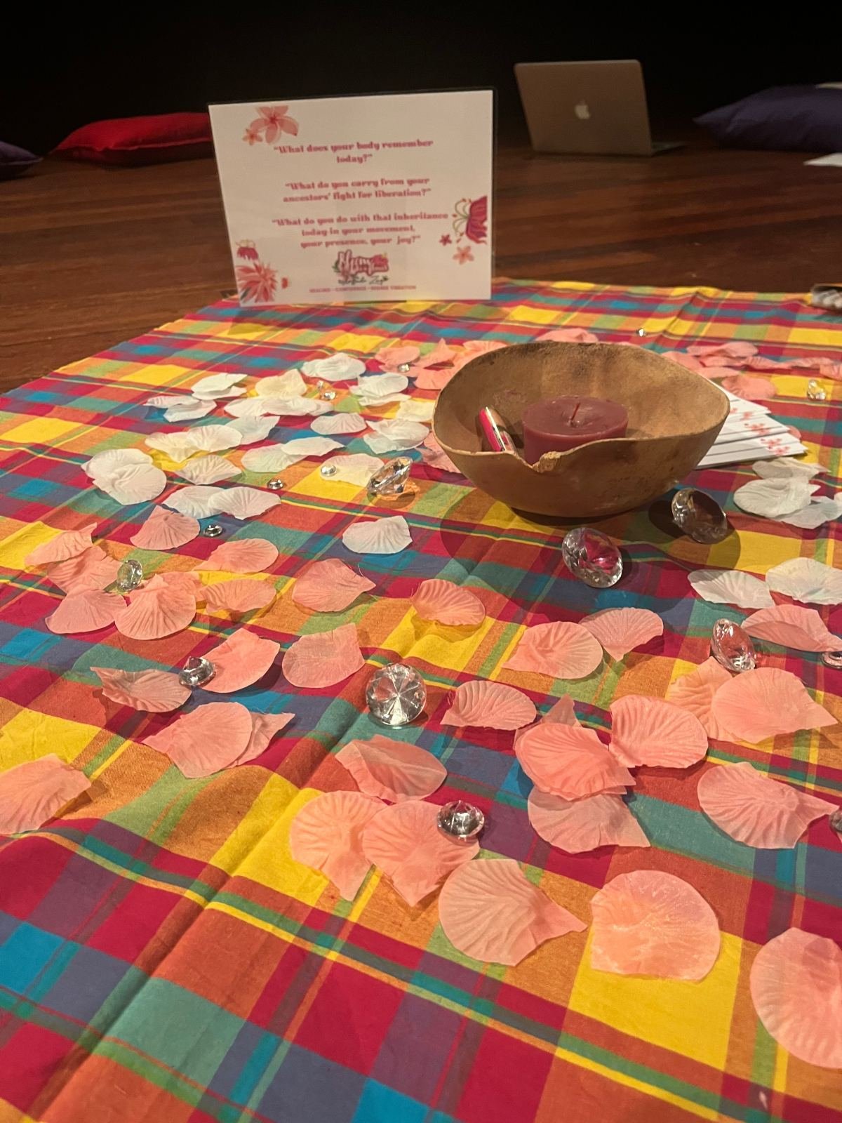 Une calebasse avec une bougie, des cristaux et des pétales roses et blancs sur un tissu madras, près d’une carte de questions à méditer